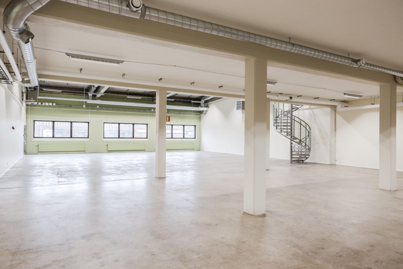 Rymlig, tom industrilokal med stora fönster, synliga kanaler, betonggolv och en spiraltrappa i metall i det bakre hörnet.