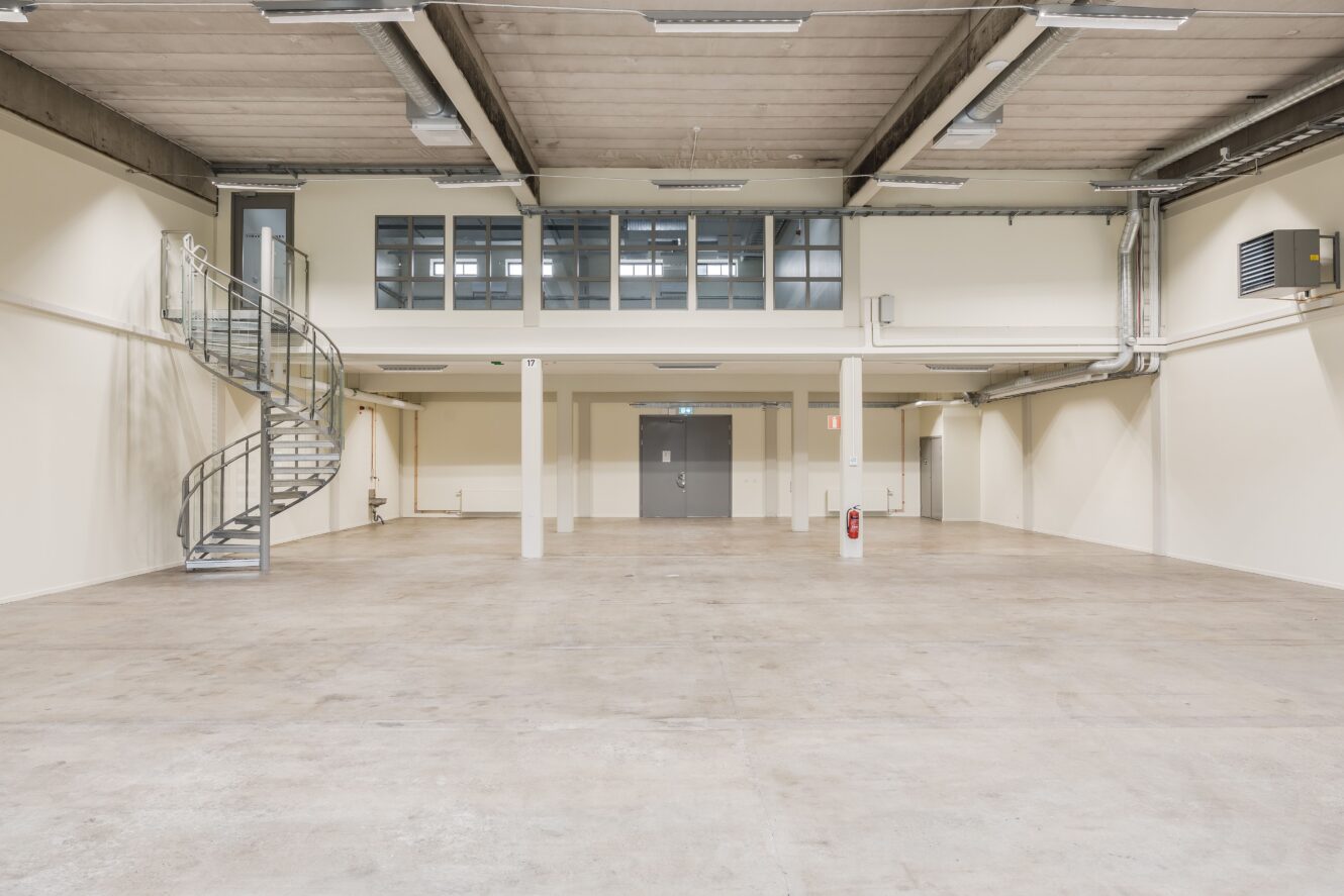 Rymlig tom industrilokal med betonggolv, entresolplan, spiraltrappa och stora dubbeldörrar under lysrörsbelysning.