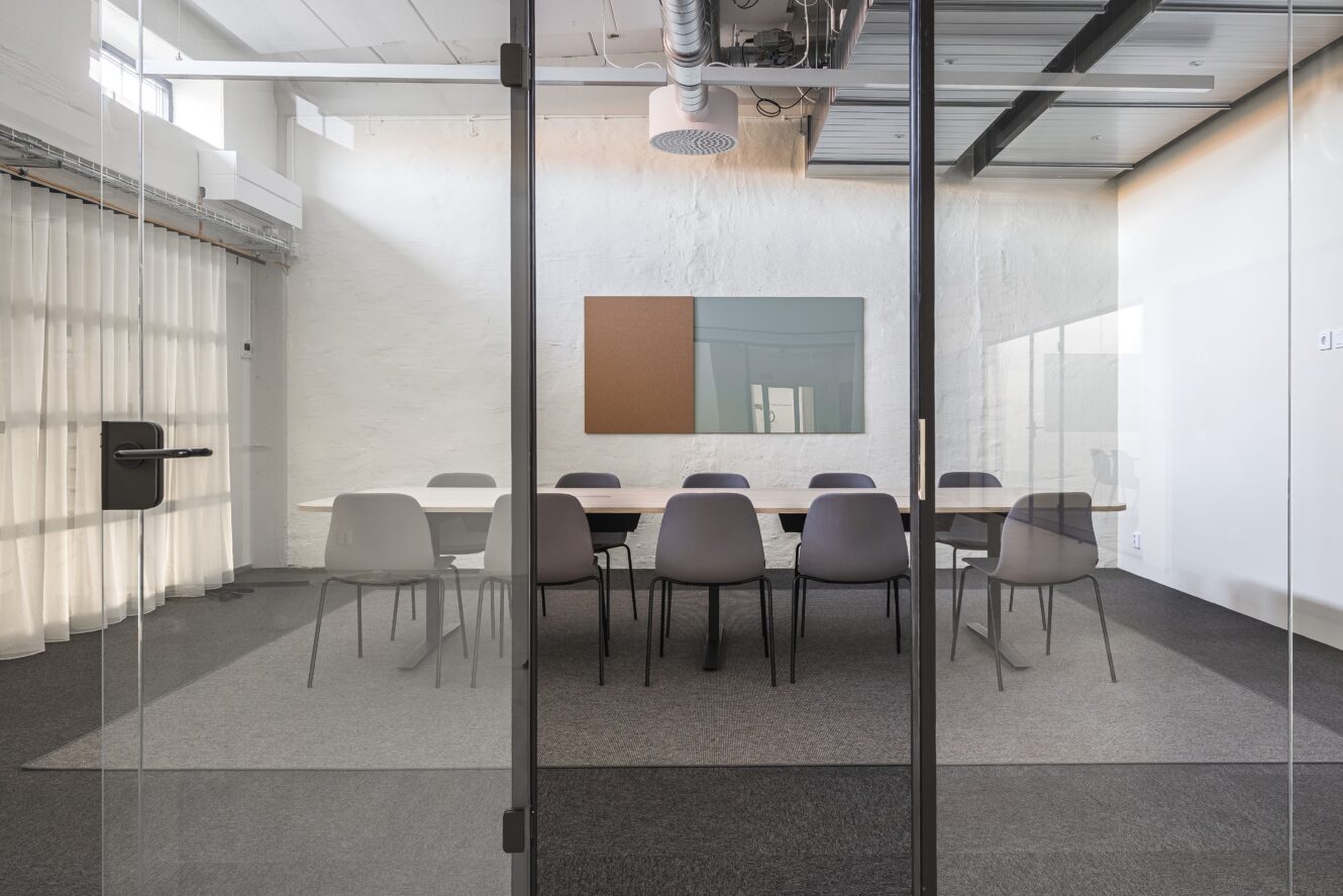 Ett modernt konferensrum med ett långt bord, tio stolar och en korktavla på väggen, sett genom glasdörrar.