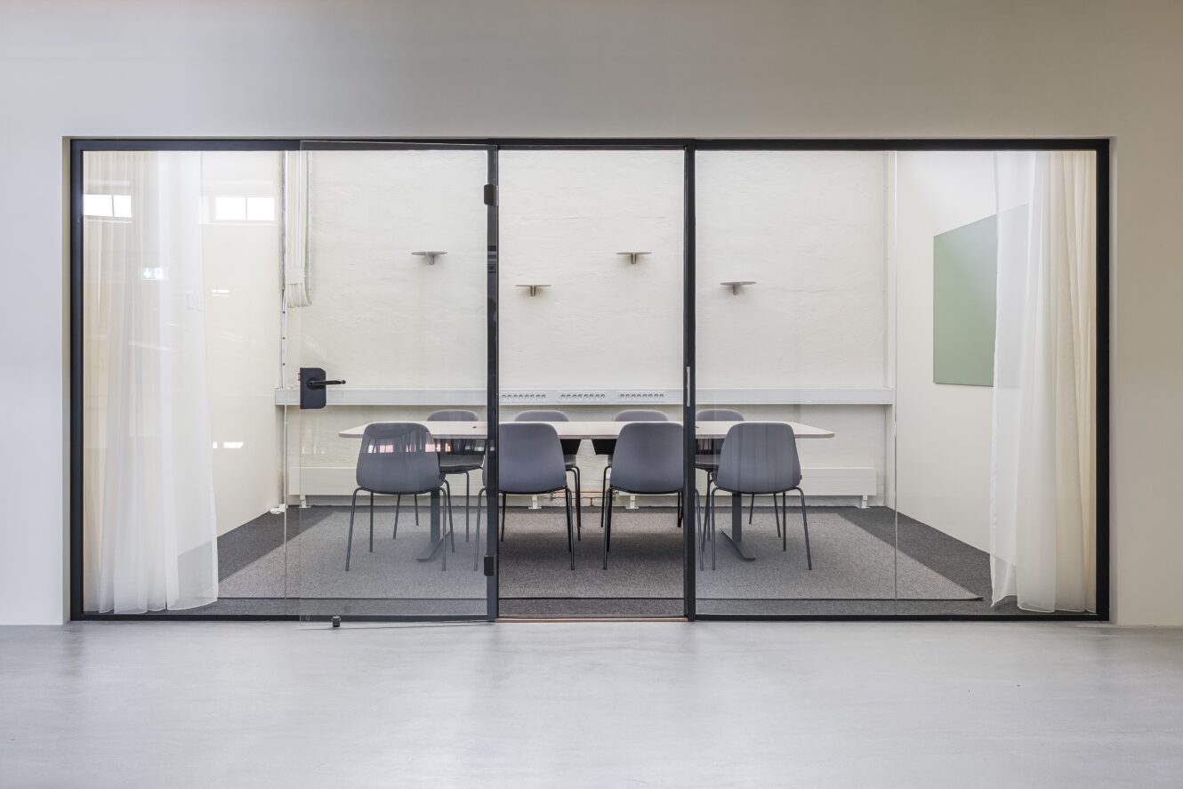 Ett modernt konferensrum med ett långt bord, åtta svarta stolar, glasväggar, vita gardiner och minimalistisk inredning.