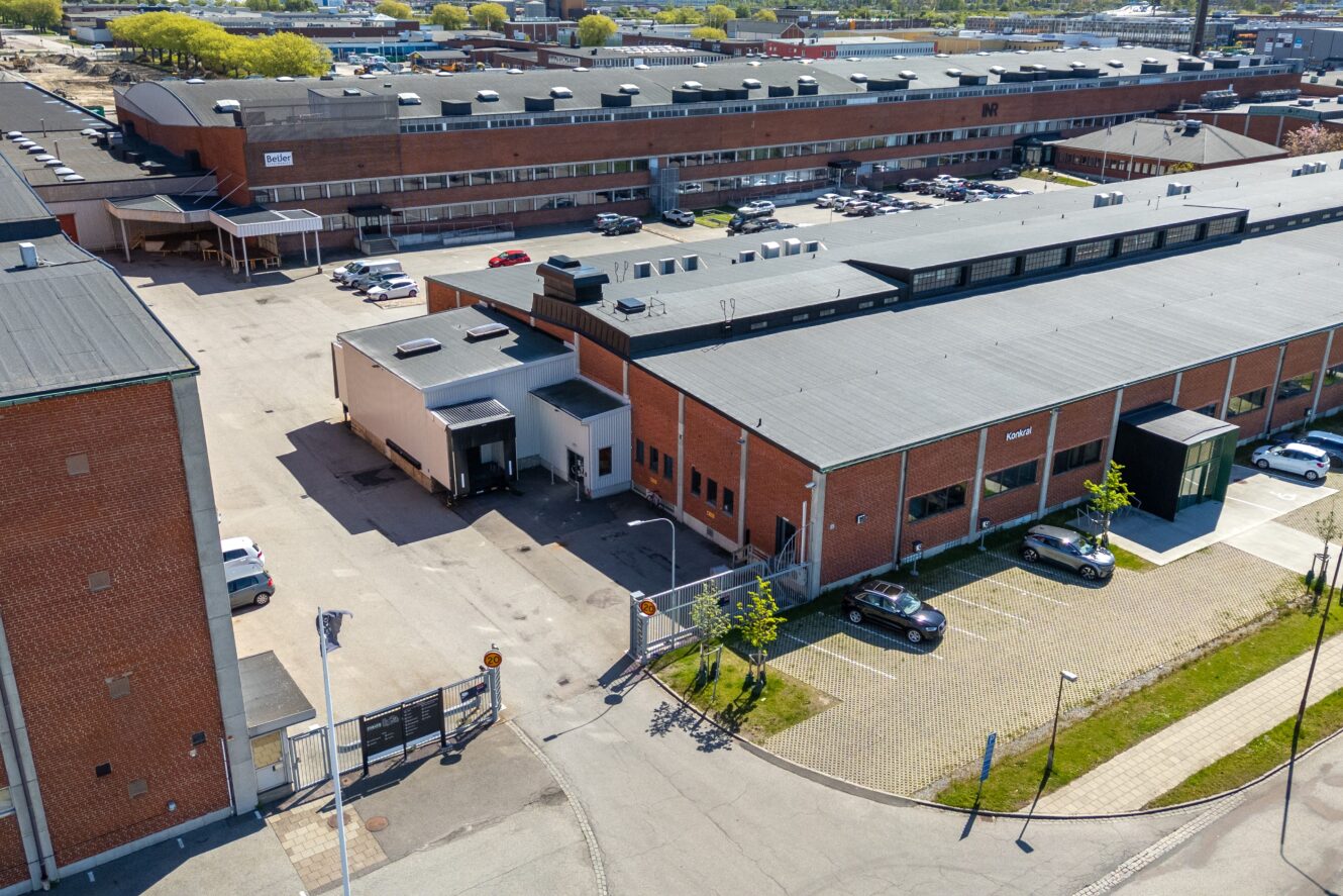 Flygfoto över ett industrikomplex med flera lagerbyggnader i tegel, parkerade bilar, lastkajer och grindförsedda entréer.