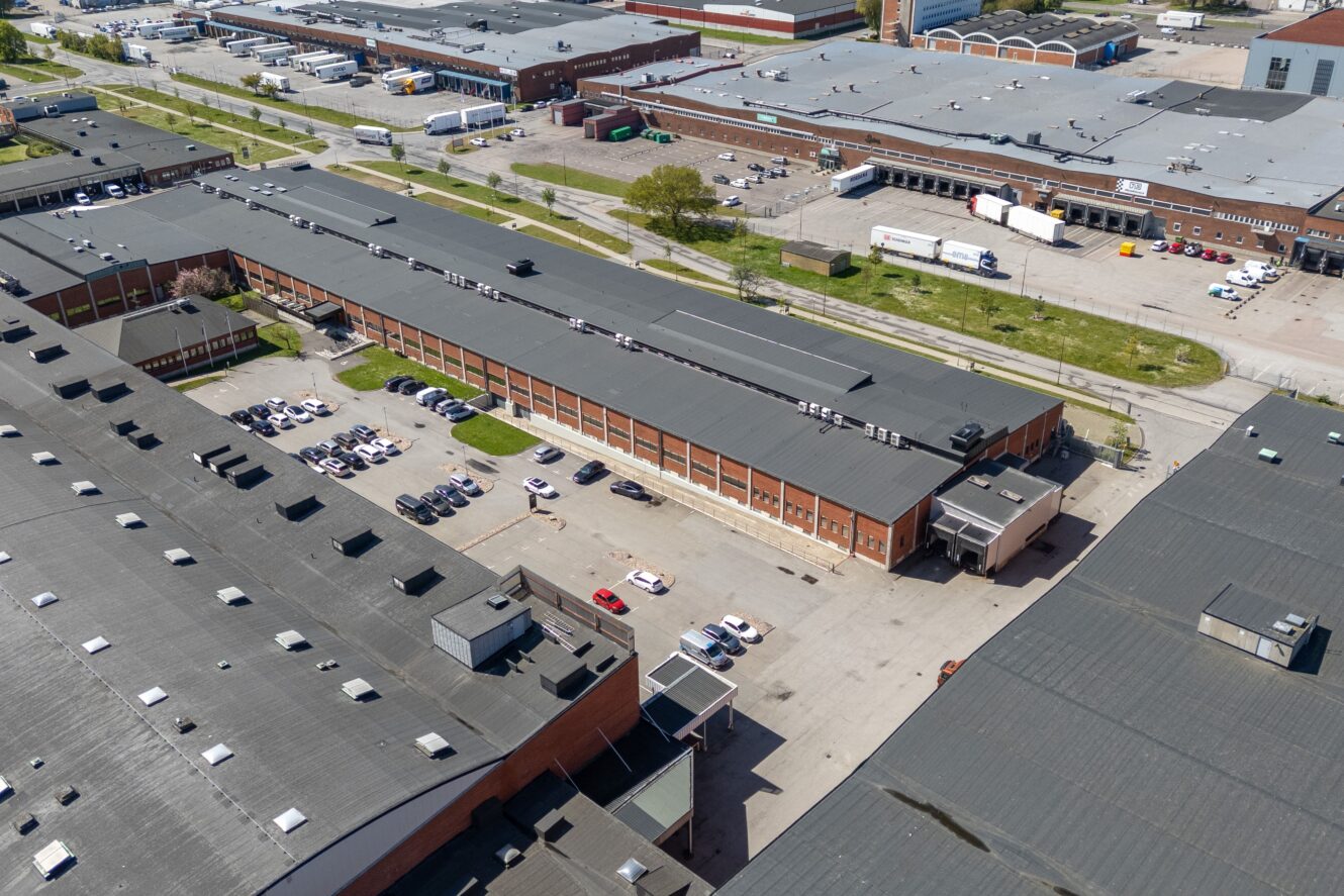 Flygfoto över ett industrikomplex med stora lagerbyggnader, parkerade bilar, lastkajer och semitrailers på parkeringsplatsen.