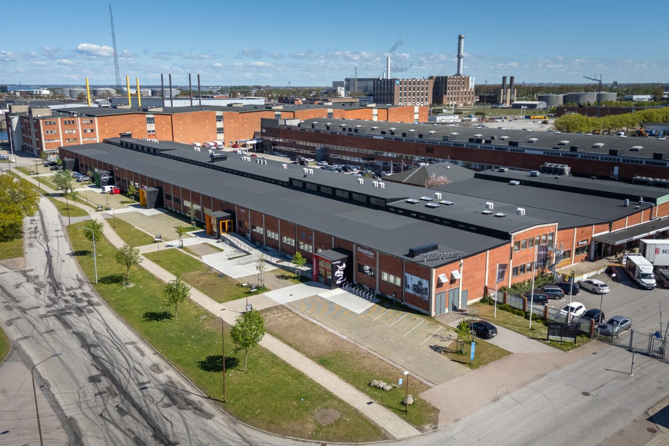 Flygfoto över ett industrikomplex med flera byggnader i rött tegel, parkeringsplatser och omgivande vägar under en klar himmel.