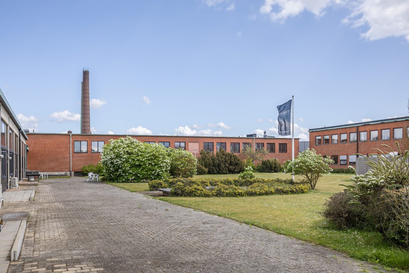 En industribyggnad i rött tegel med många fönster vetter mot en anlagd innergård med gröna buskar, blommor och en flaggstång under en blå himmel.