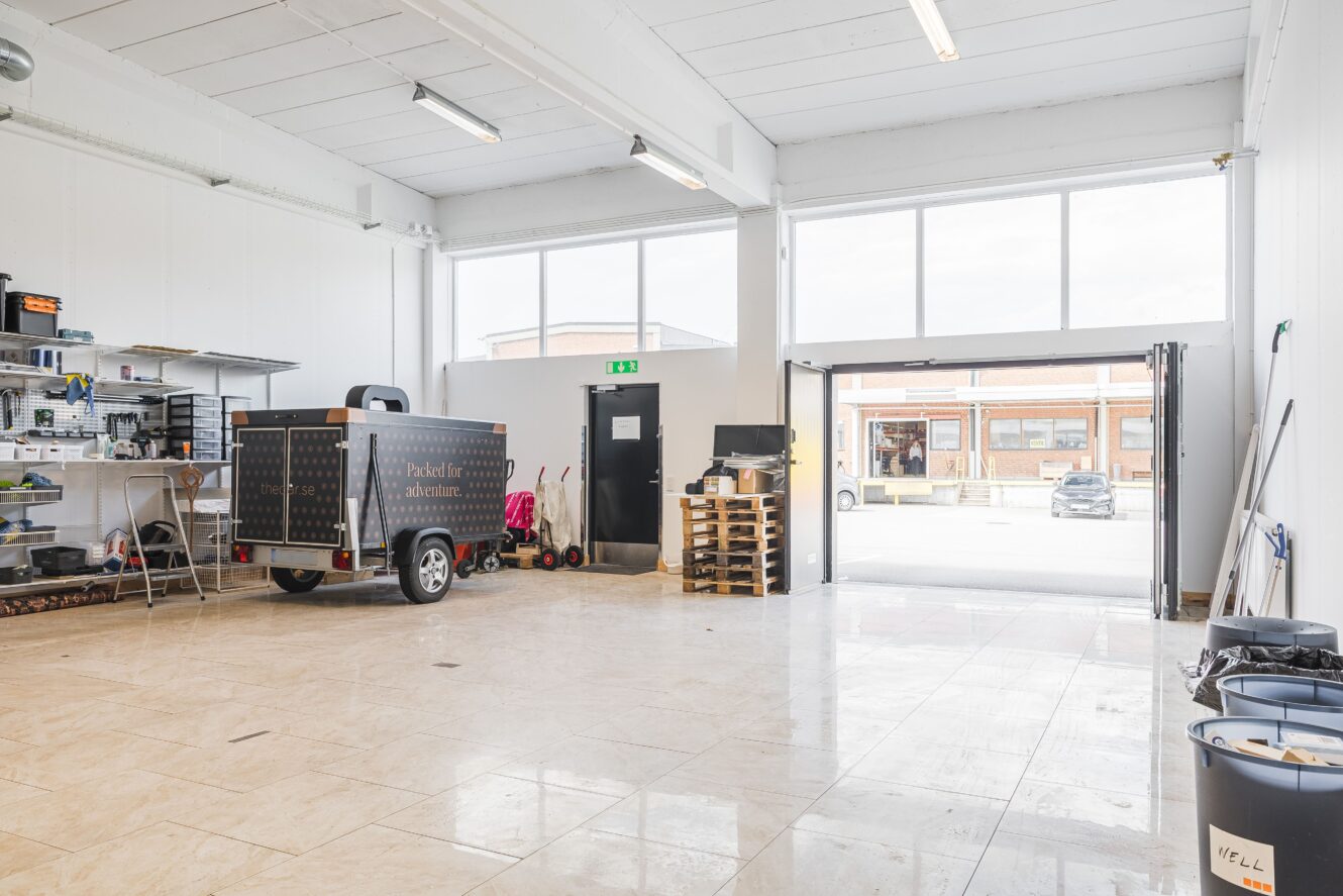 Ett rymligt, rent garage med hyllor, en släpvagn, städutrustning, pallar och en öppen garageport med bilar och byggnader utanför.