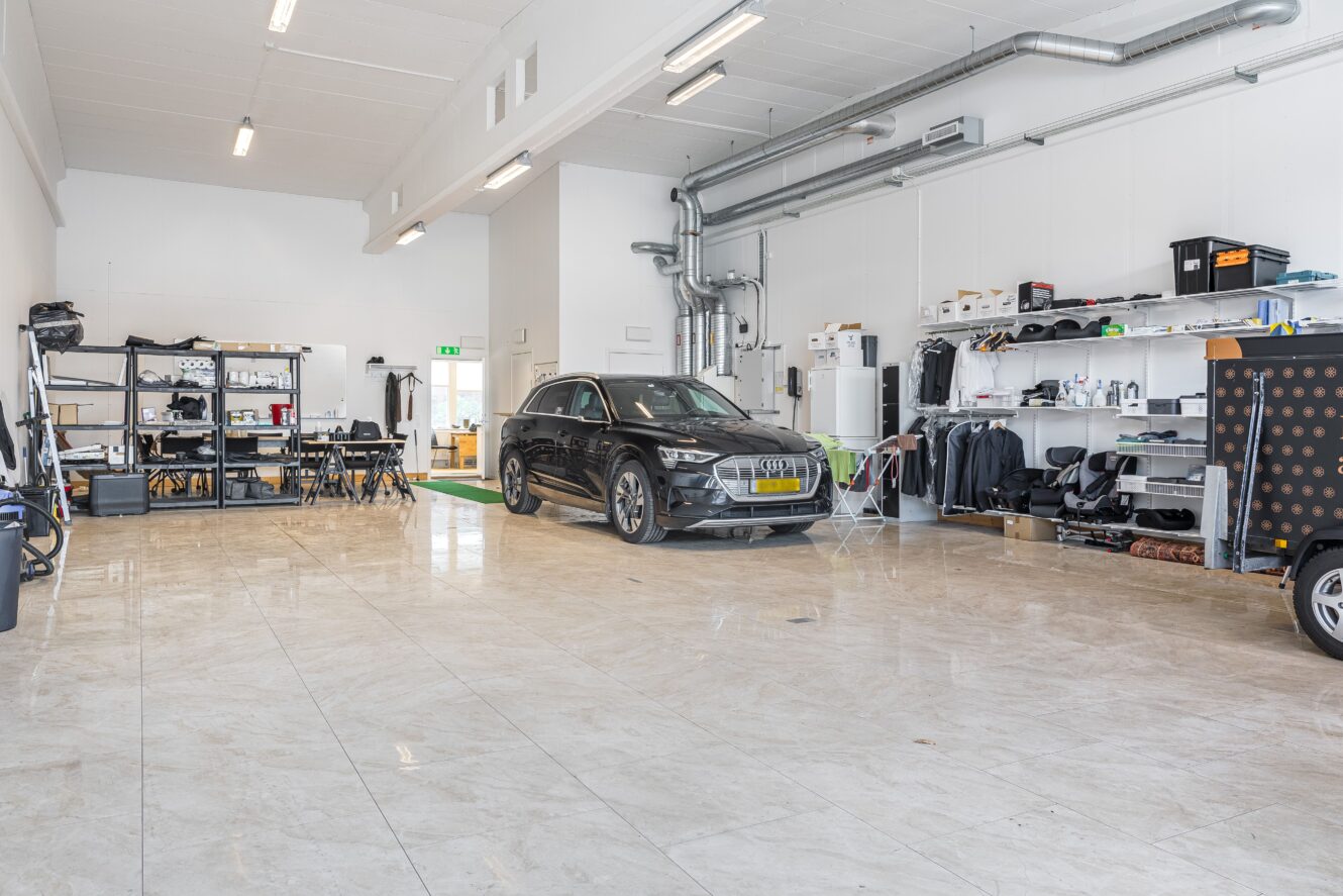 En svart bil står parkerad i ett rymligt, väl upplyst garage med hyllor och utrustning längs väggarna.