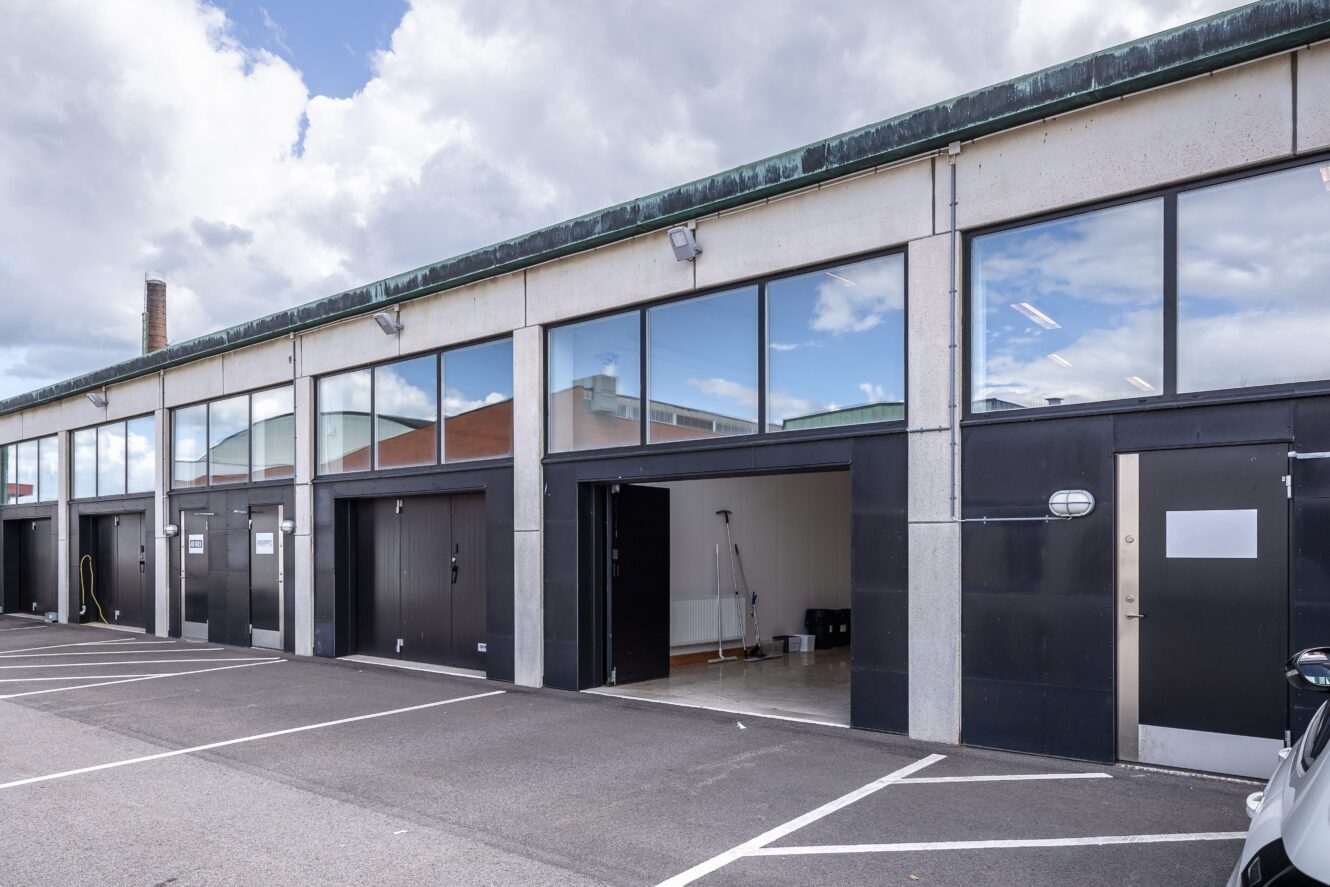 En rad moderna garageenheter med stora fönster ovanför svarta dörrar; en garageport är öppen och avslöjar rengöringsutrustning inuti. Området framför är en tom parkeringsplats.