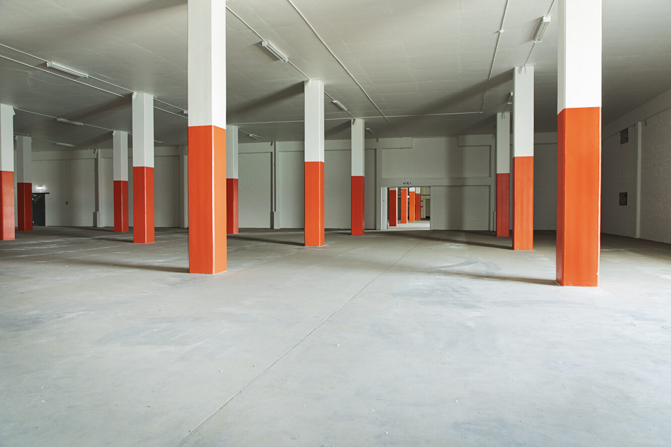 Stor tom lagerlokal med betonggolv, många vita pelare med orange bas och fluorescerande belysning i taket.