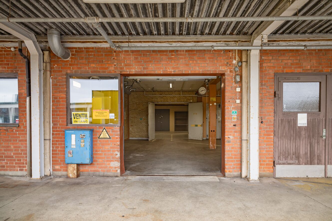 En öppen garageport i en industribyggnad i tegel med en blå varuautomat, gul skyltning och synliga rör i taket.