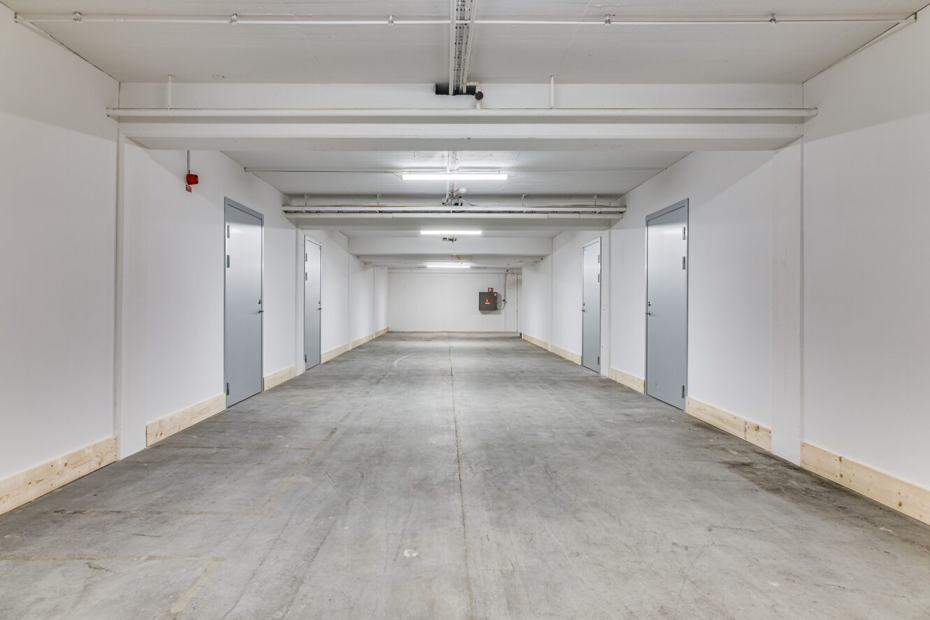 En ren, tom underjordisk korridor med betonggolv, vita väggar och flera stängda grå dörrar på båda sidor. Rör och lampor i taket är synliga.