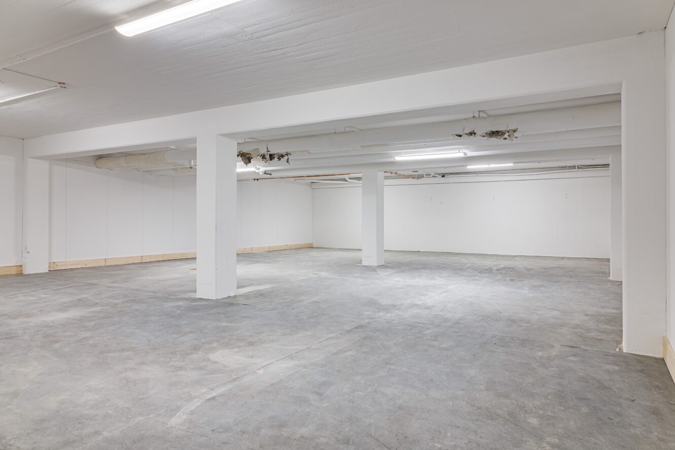 En tom, oinredd källare med betonggolv, vita väggar, synliga rör i taket och lysrörsbelysning.