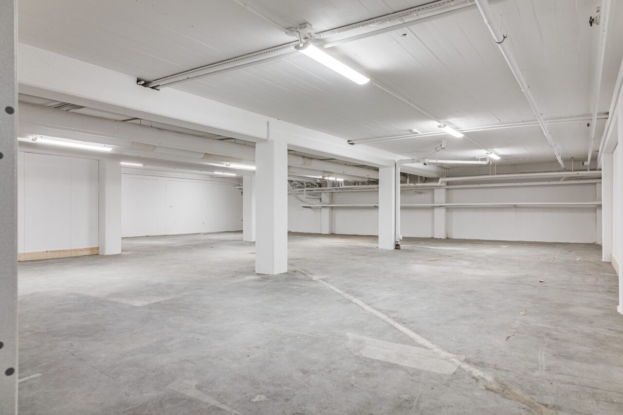 Stort tomt källarutrymme med betonggolv, vita väggar, stödpelare och lysrörsbelysning.
