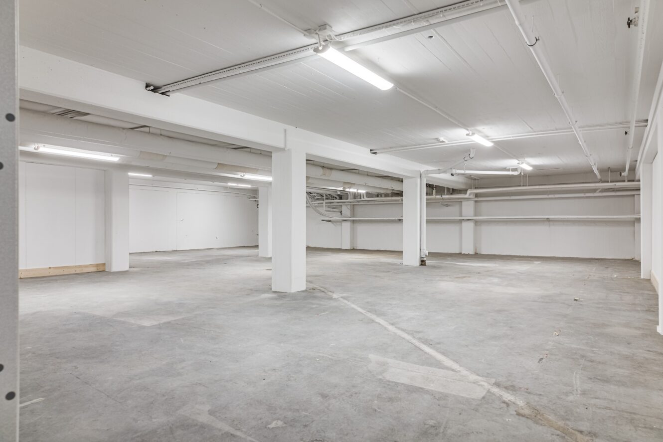 Stort tomt källarutrymme med betonggolv, vita väggar, stödpelare och lysrörsbelysning.