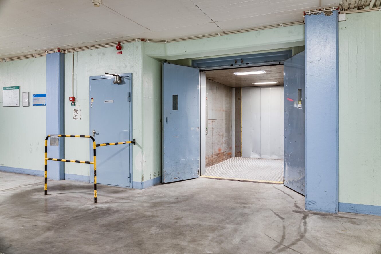 Godshiss med öppna dörrar i ett industriellt källarområde med betonggolv och en säkerhetsbarriär av metall i närheten.