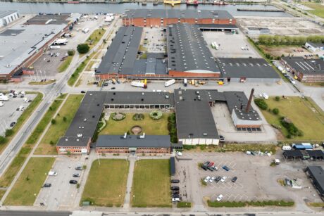 Flygfoto över ett industrikomplex med flera stora lagerbyggnader, parkerade lastbilar, bilar och gröna gräsmattor, nära en strandpromenad.