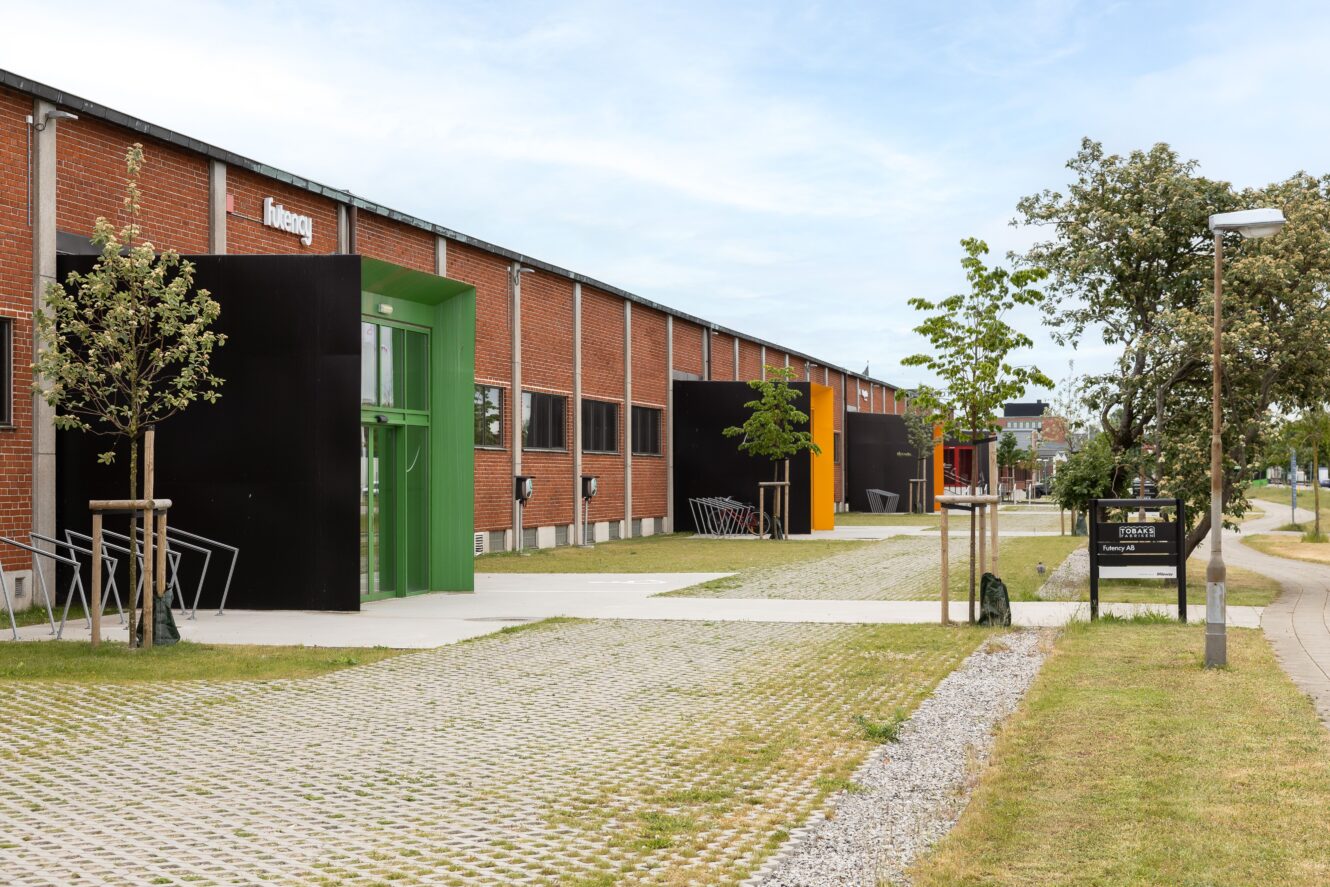 En modern tegelbyggnad med färgglada dörrkarmar, unga träd, cykelställ och en skylt med texten Forsker Parken längs en asfalterad gångväg och gräsyta.