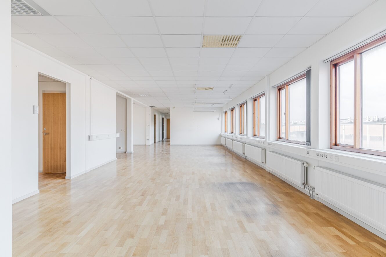 Tomt kontorsutrymme med ljust trägolv, vita väggar, takplattor och stora fönster längs ena sidan som släpper in naturligt ljus.