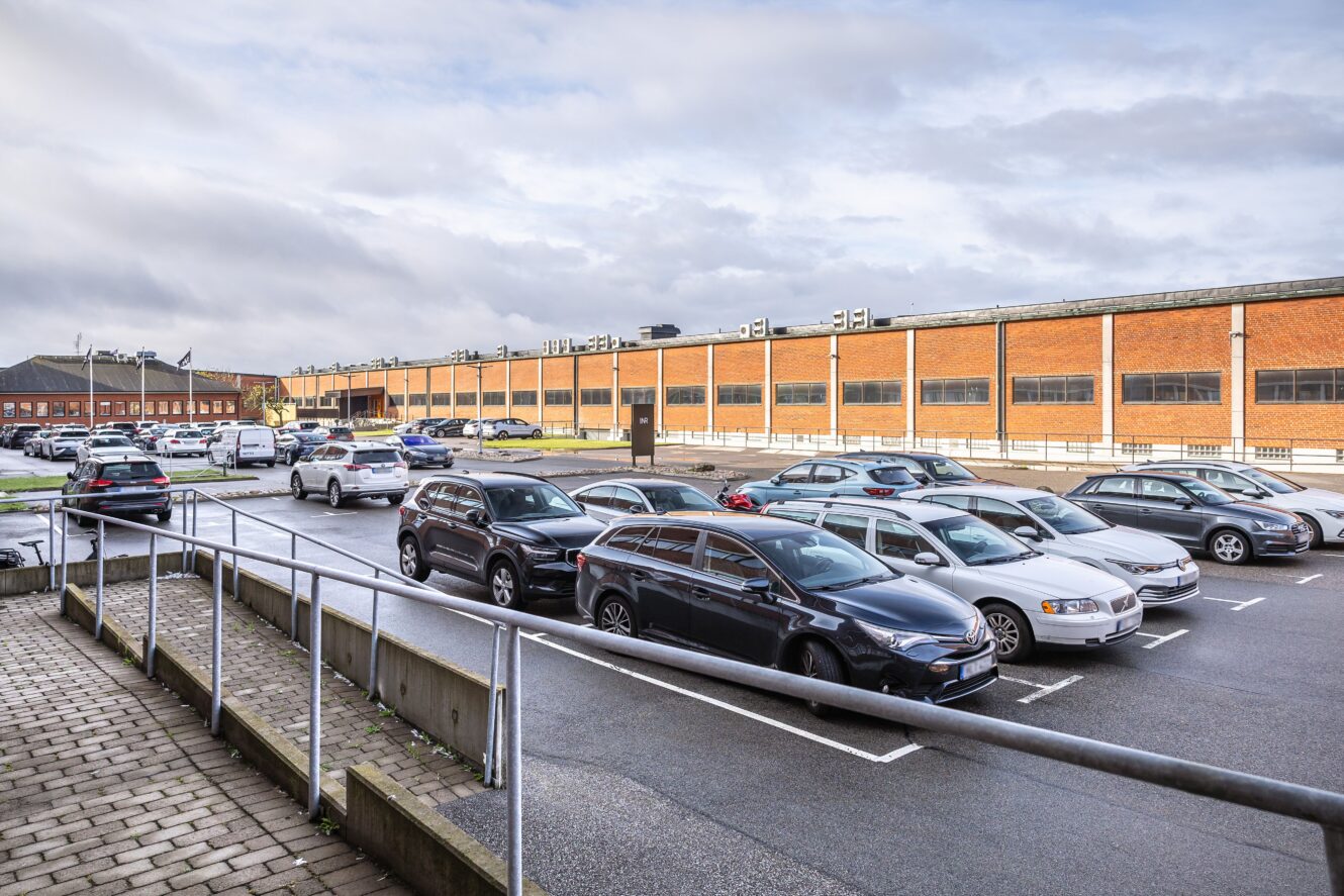 En parkeringsplats med flera parkerade bilar framför en lång industri- eller handelsbyggnad i tegel under en molnig himmel.