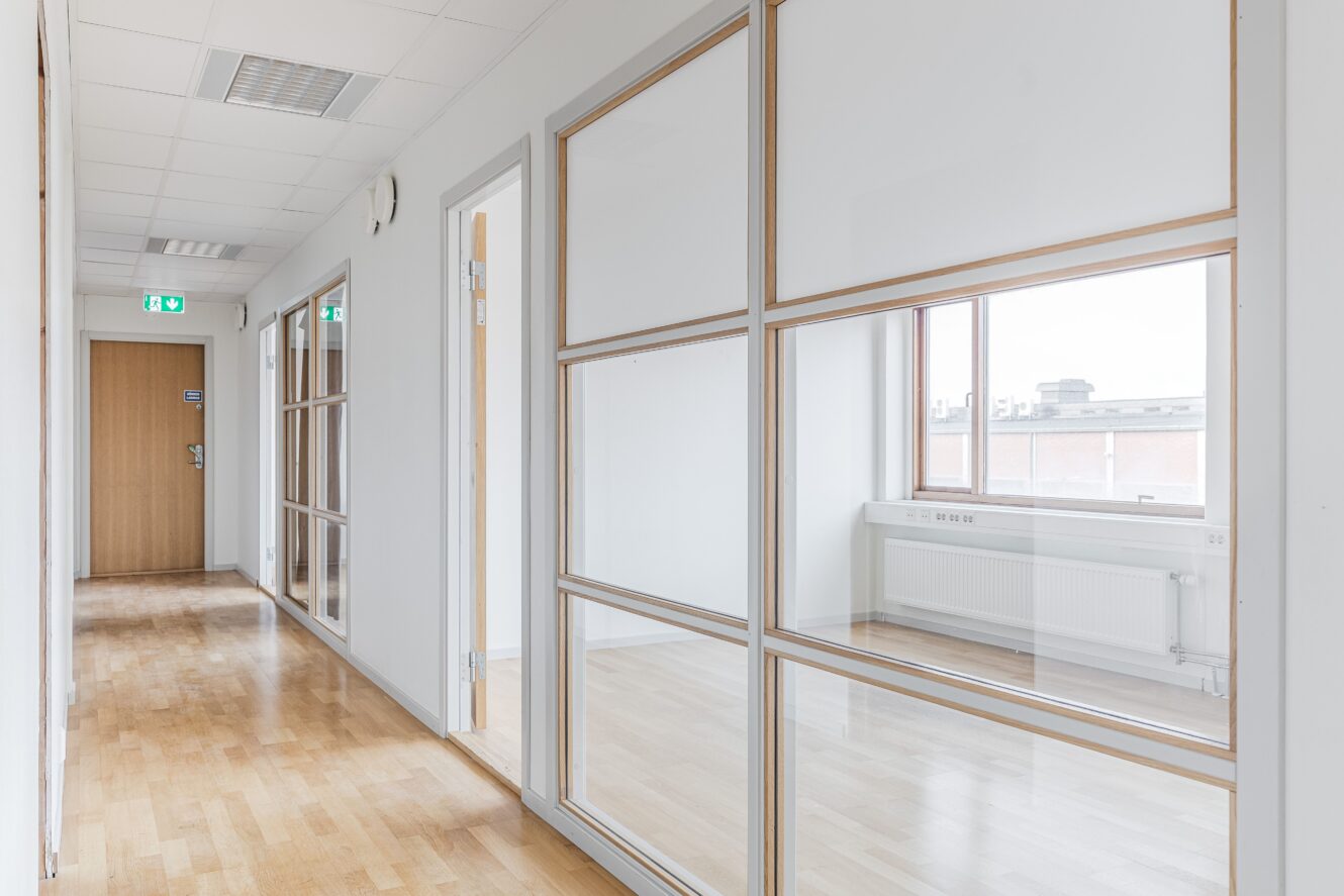 En modern kontorskorridor med ljusa trägolv, vita väggar, glaspartier och en trädörr i slutet.