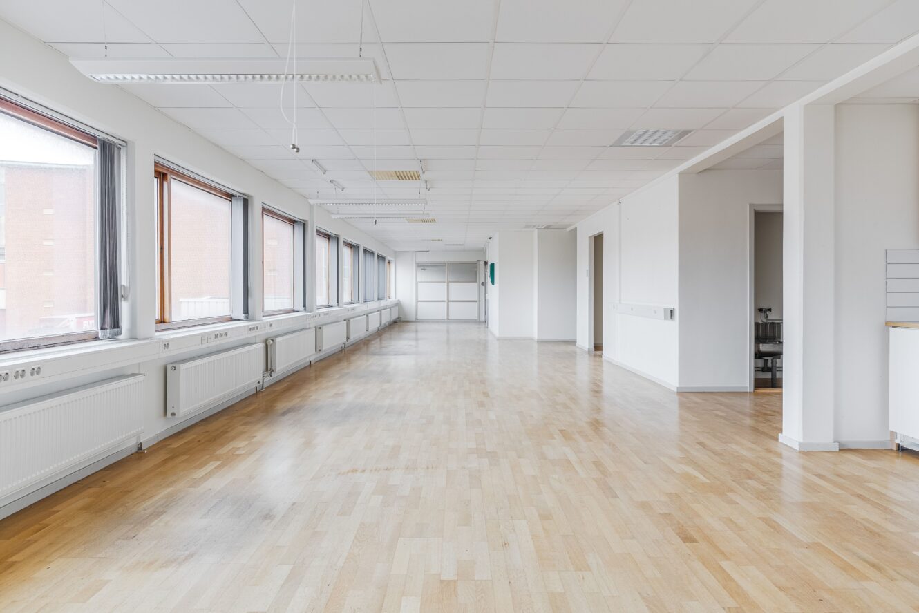 Stort, tomt kontorsutrymme med ljust trägolv, vita väggar, en rad fönster till vänster och taklampor.