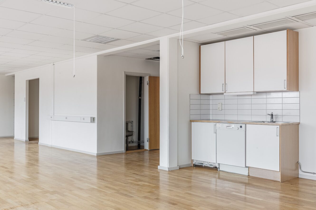 Ett tomt rum med ljust trägolv, vita väggar, ett litet pentry med skåp, diskho och kaklat stänkskydd. Taklampor och eluttag är synliga.