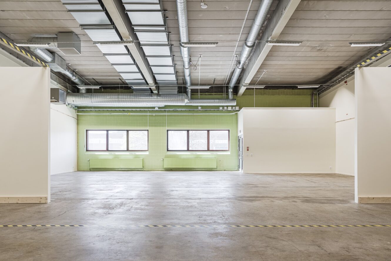 En rymlig, tom industrilokal med synliga takkanaler, gröna kaklade väggar, stora fönster och ett betonggolv med randig tejp.