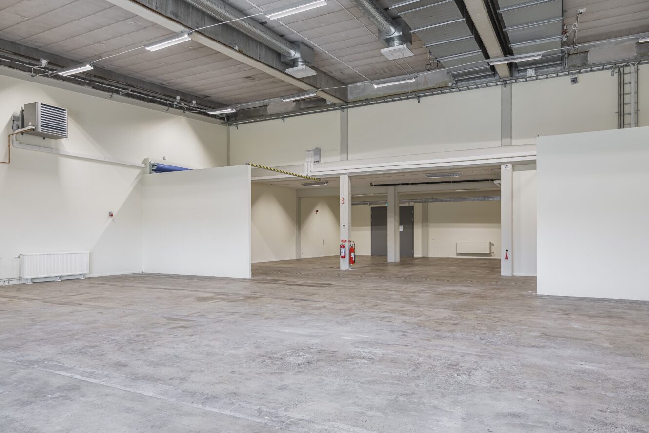 Rymlig, tom industri- eller lagerlokal med vita väggar, betonggolv, takbelysning och synliga ventilationskanaler. Brandsläckare är monterade på stödpelare.