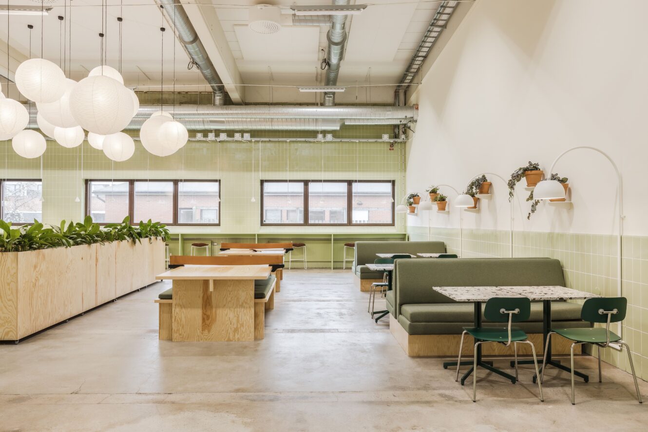Modern, minimalistisk cafeteria med gröna bås, träbord, hängande glober, krukväxter och stora fönster som släpper in naturligt ljus.