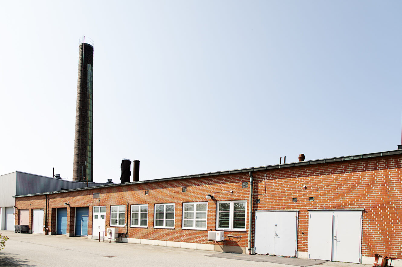 En envånings industribyggnad i rött tegel med flera dörrar, fönster och en hög skorsten under en klar himmel.