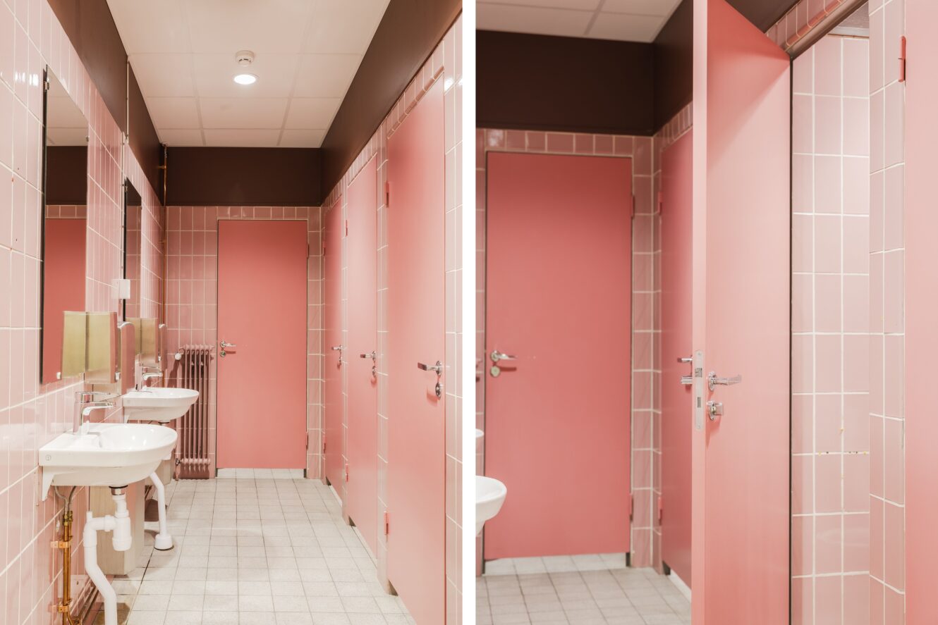 En offentlig toalett med rosa kaklade väggar, rosa dörrar, två vita handfat, speglar och ett kaklat golv under taklampor.