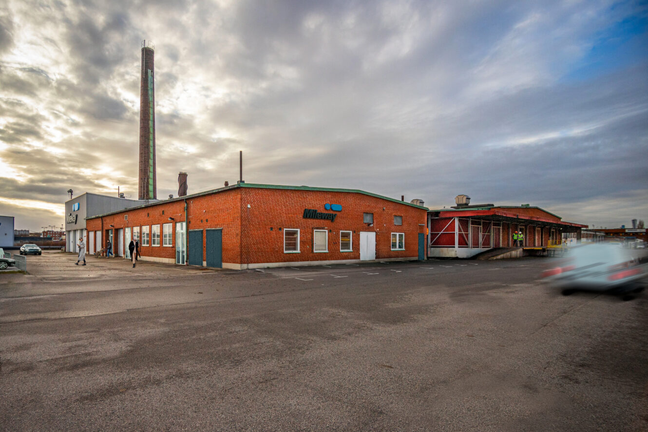 En industribyggnad i rött tegel med en hög skorsten under en molnig himmel; några människor går i närheten och en oskarp bil rör sig på parkeringsplatsen.