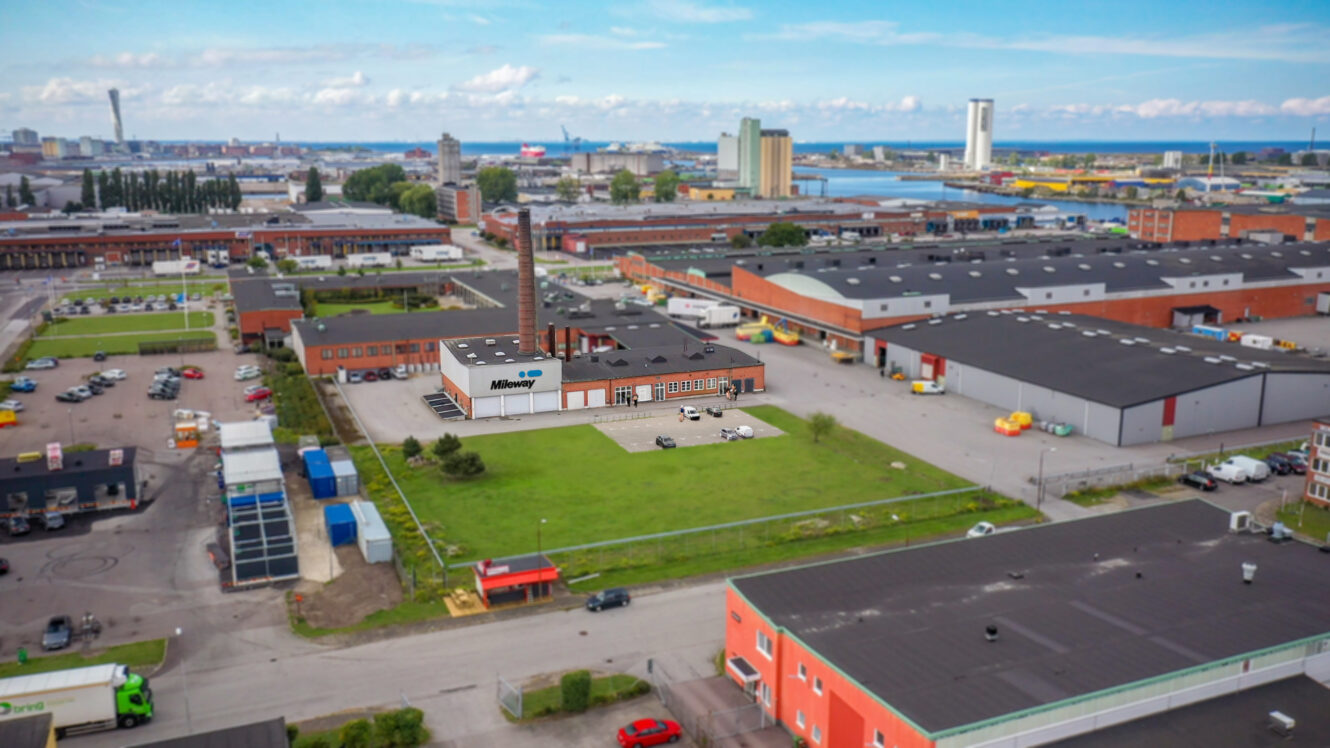 Flygfoto över ett industriområde med lagerbyggnader, parkeringsplatser och ett gräsbevuxet fält under en blå himmel med spridda moln.