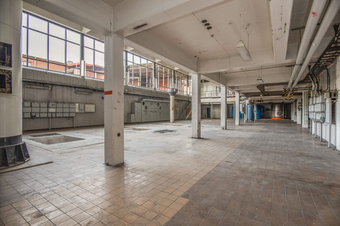 Rymlig, tom industriinredning med klinkergolv, stora fönster och synliga rör; viss väggmonterad utrustning och en öppen grop syns.