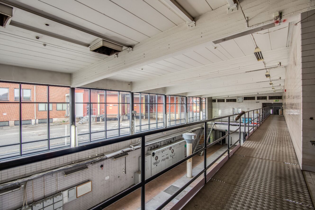 Invändig vy av en industribyggnad med stora fönster, metallräcken, kaklade väggar och en gångväg med utsikt över lägre nivåer.