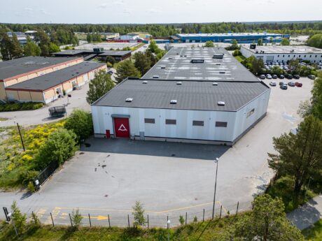Ett stort vitt industriellt lager med platt tak och röd entrédörr, omgivet av asfalterat område, träd och närliggande kommersiella byggnader.