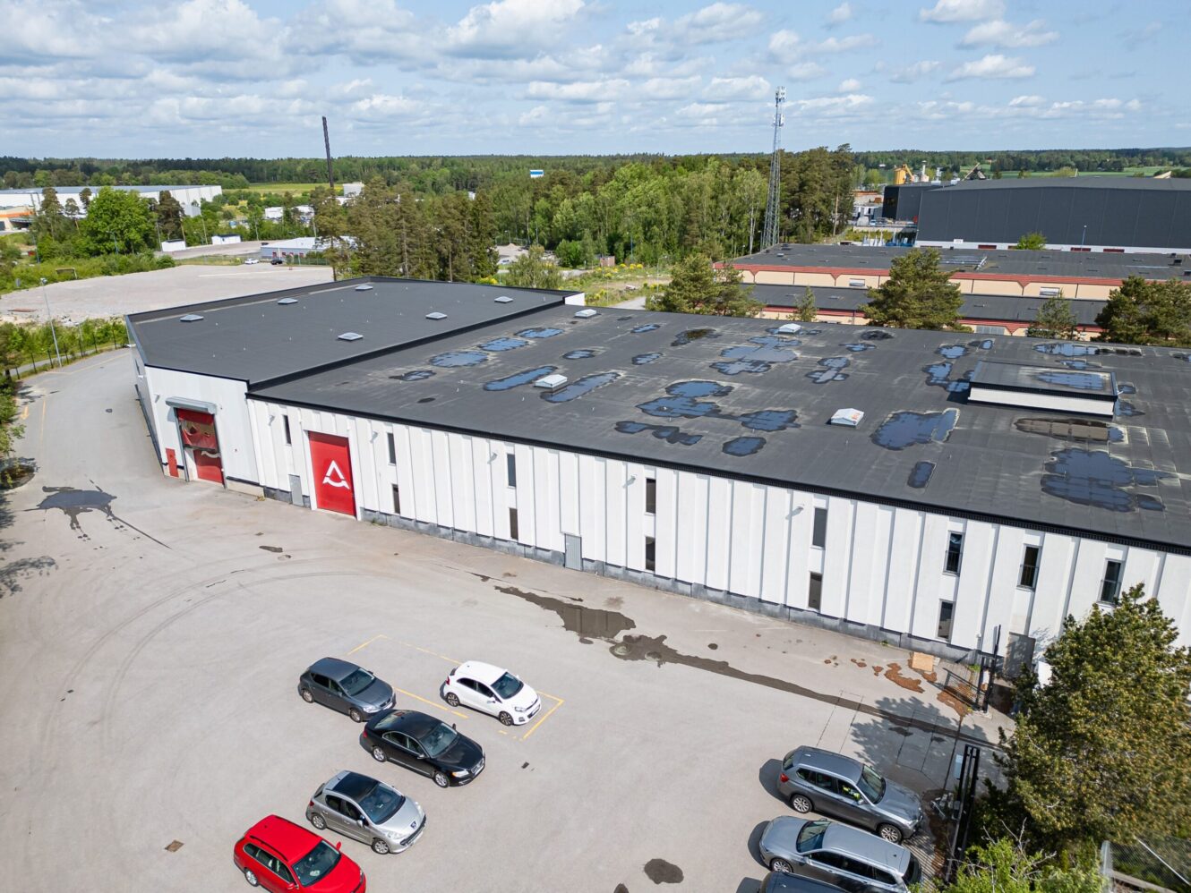 Flygfoto över en stor vit industrihall med flera bilar parkerade på parkeringen och vattenfläckar på taket, omgiven av träd och andra industribyggnader.