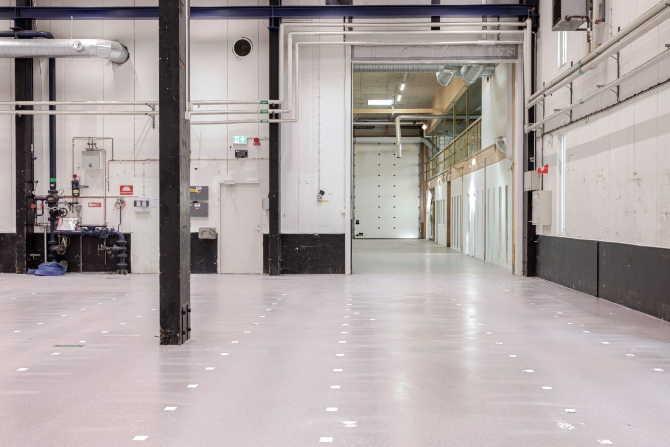 Stort tomt industrilager med polerade golv, högt i tak, synliga rör och en rad vita dörrar längs ena väggen.