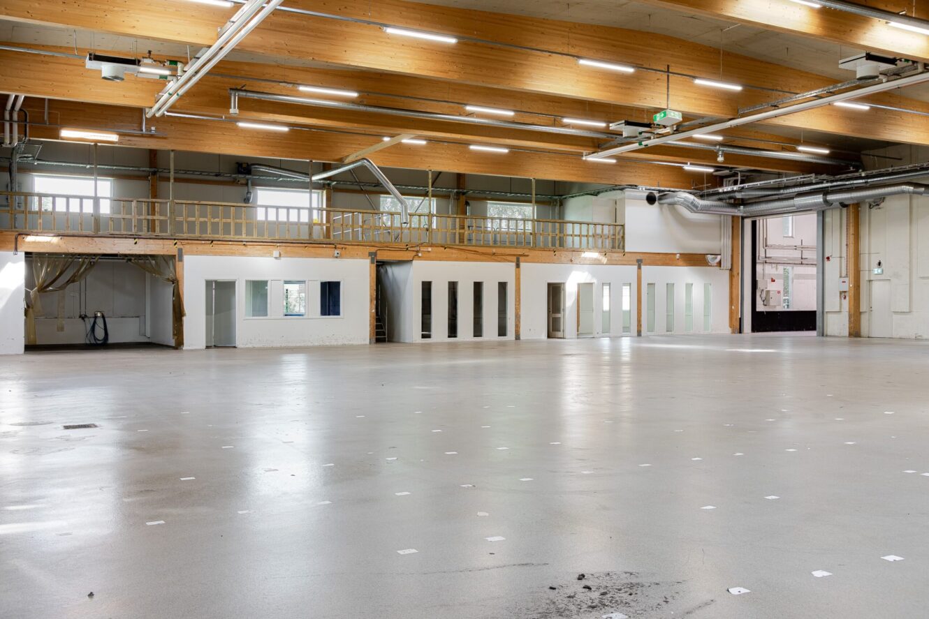Rymlig, tom industrilokal med högt i tak, synliga bjälkar, takbelysning och en mezzaninvåning längs bakväggen.