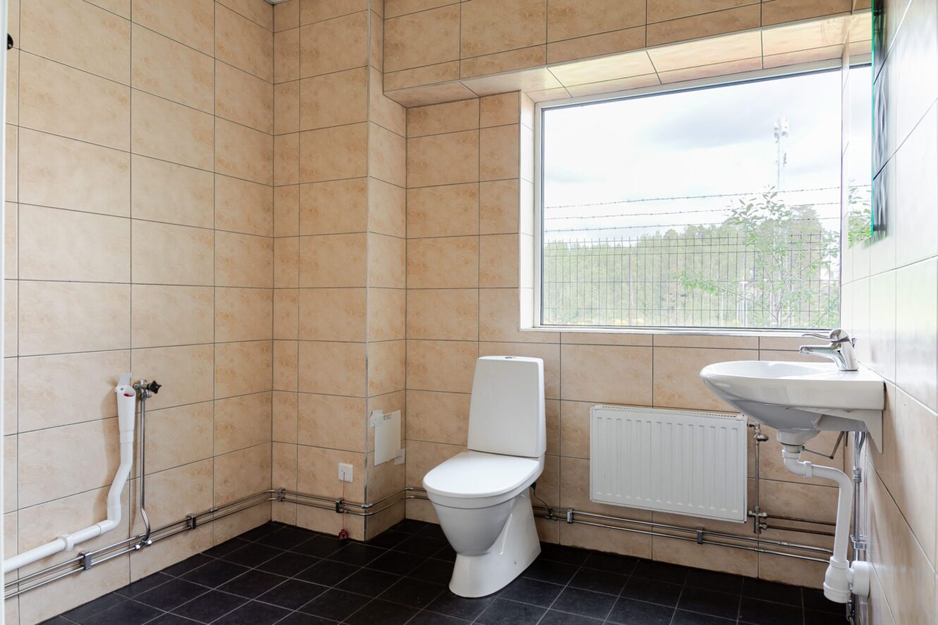 Ett rent, kaklat badrum med toalett, väggmonterat handfat, radiator, vattenarmaturer och ett stort fönster som släpper in naturligt ljus.