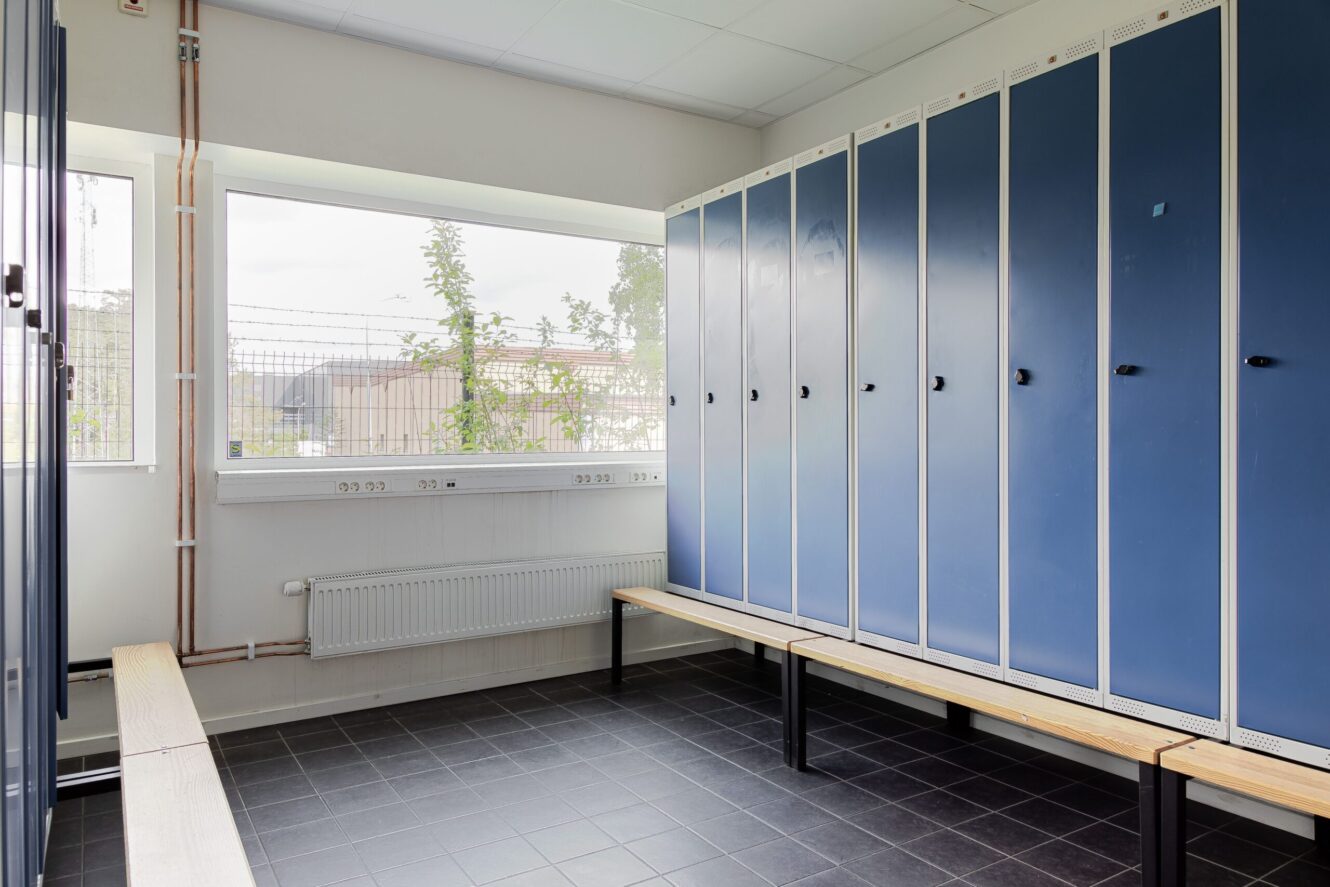 Ett rent omklädningsrum med blå skåp, ljusa bänkar, klinkergolv och stora fönster som släpper in naturligt ljus.