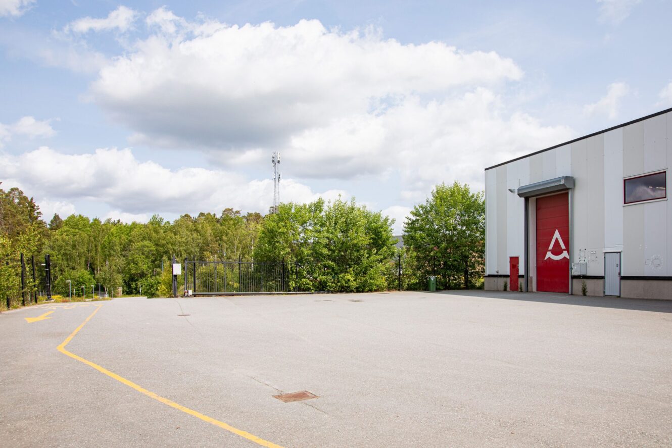 En tom industritomt med en gul pil på asfalten, bredvid en vit byggnad med en röd garageport och träd i bakgrunden under en delvis molnig himmel.