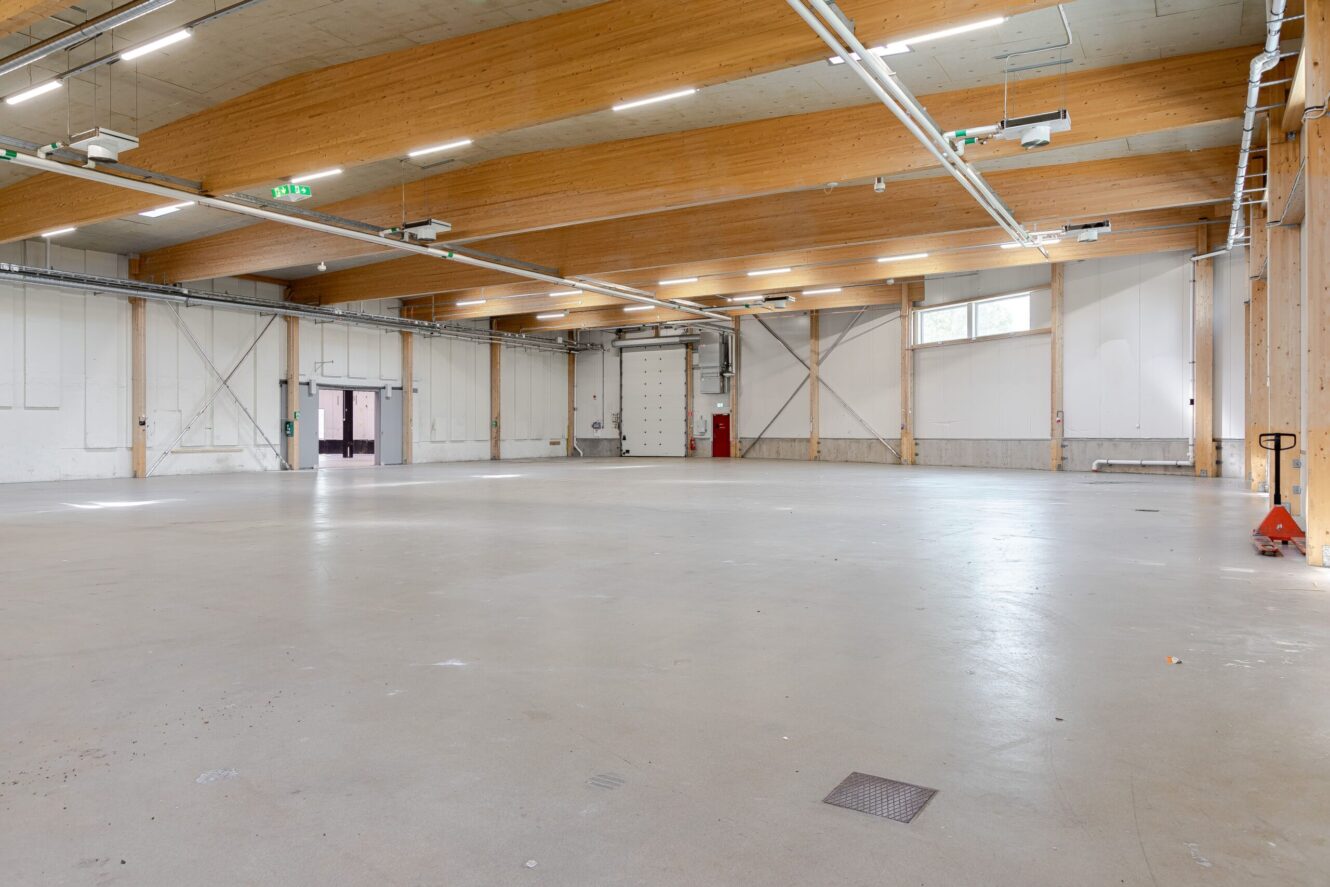 Rymlig, tom lagerlokal med betonggolv, högt trätak, stor rullport, lysrörsbelysning och minimal utrustning.