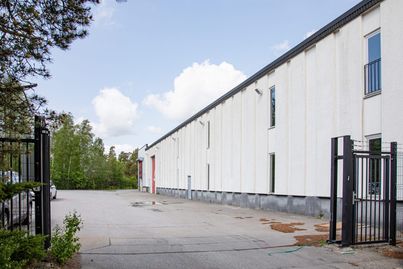 En lång vit industribyggnad med flera fönster och ett svart staket, omgiven av träd och en asfalterad tomt under en delvis molnig himmel.