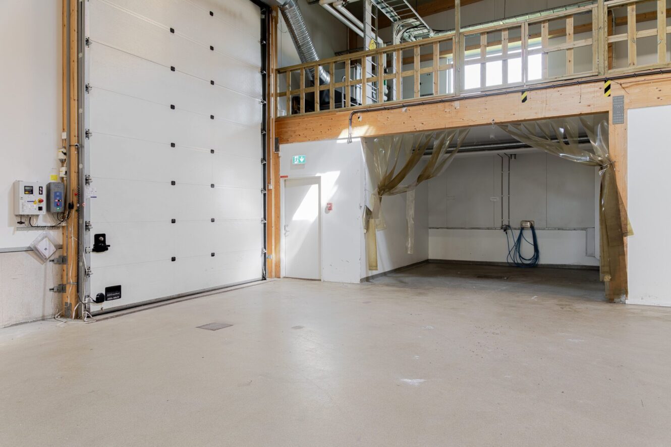 Ett tomt industriellt lagerutrymme med en stor garageport, högt i tak, loft med räcke och en liten sluten del under loftet.