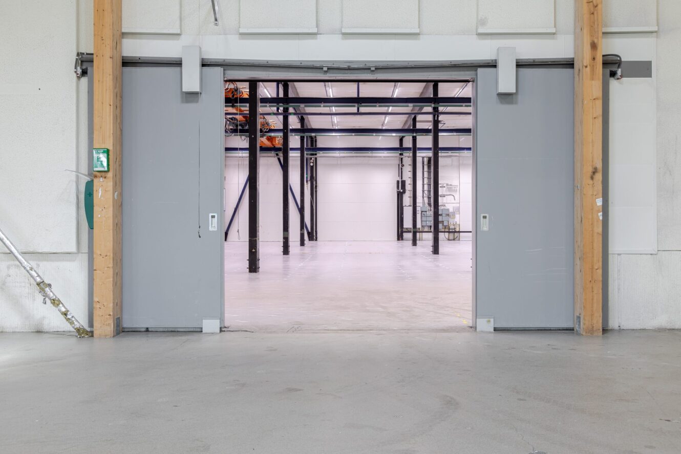 Breda skjutdörrar öppnas och avslöjar ett tomt industrilager med vita väggar, synliga bjälkar och ett polerat betonggolv.