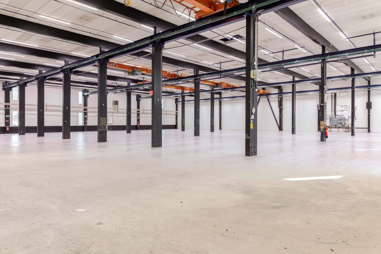Rymlig, tom lagerlokal med synliga stålbalkar, lyftkranar och ett polerat betonggolv.