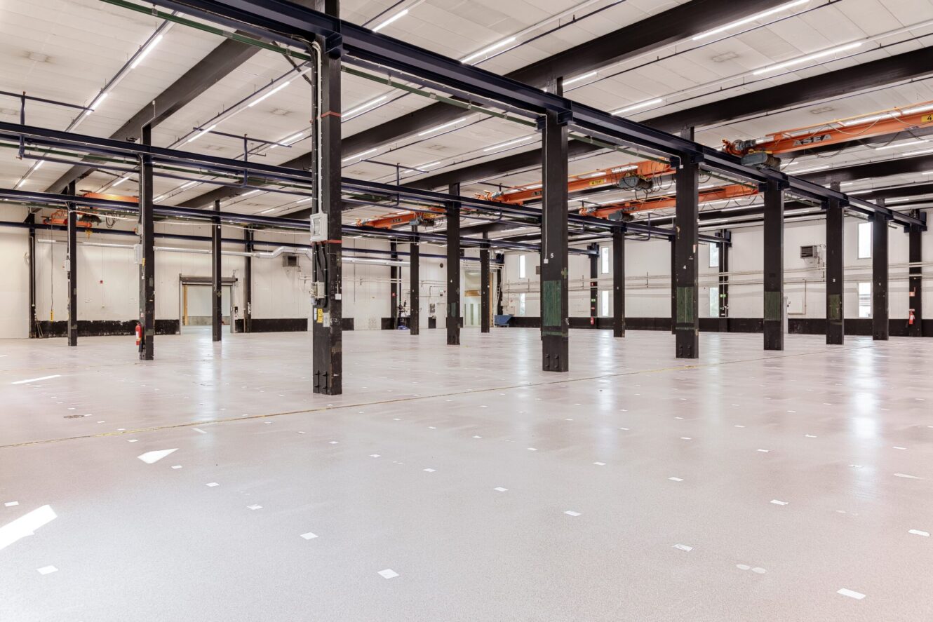 Rymlig, tom industrilagerlokal med högt i tak, stora stödbalkar, rörledningar och polerade betonggolv.