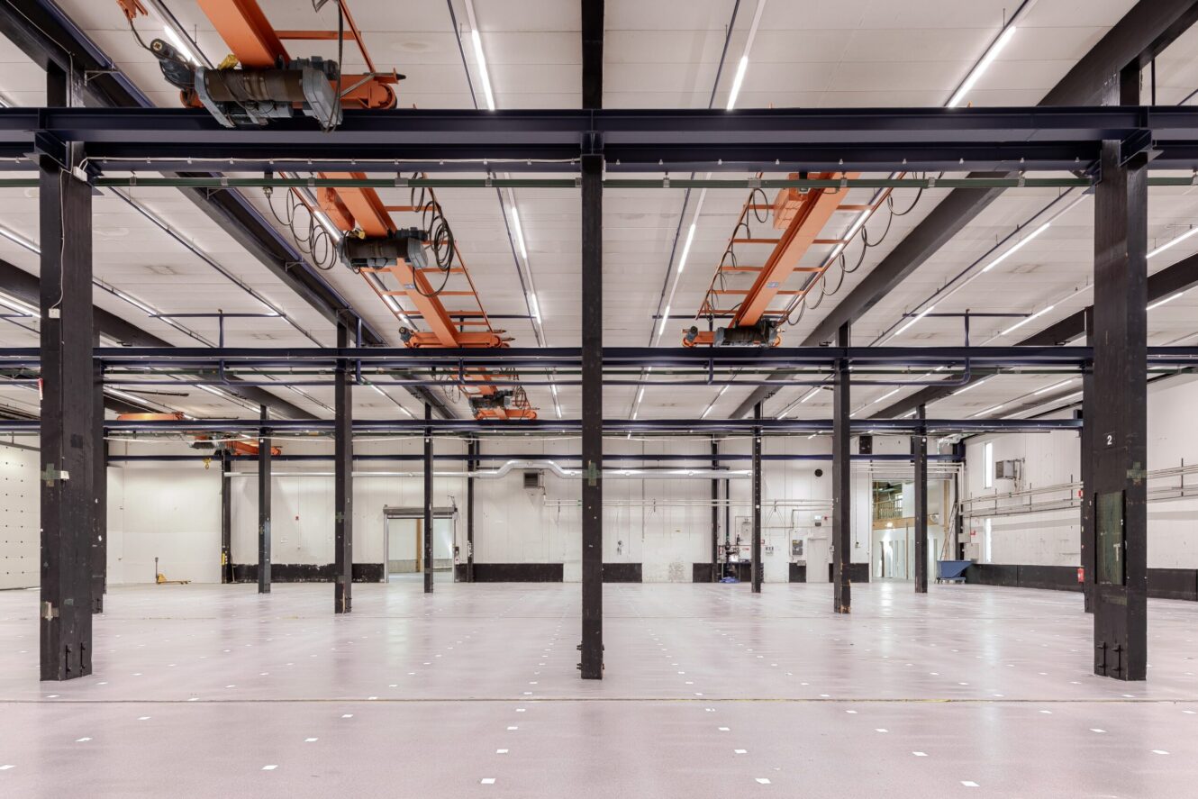 Rymlig, tom industrihall med högt i tak, lyftkranar, synliga bjälkar och polerade golv.
