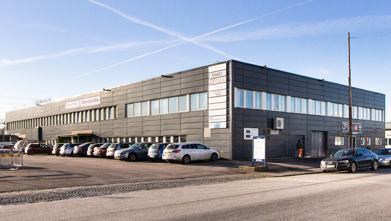 En stor, modern industribyggnad i två våningar med flera parkerade bilar framför under en klarblå himmel.