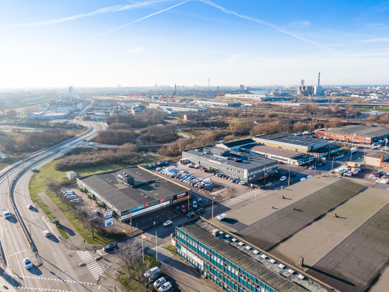 Flygbild över ett industriområde med lagerlokaler, kontorsbyggnader, parkeringsplatser, vägar och gles grönska under en klarblå himmel.